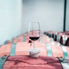 Copa de vino tinto sobre un barril de madera, con una bodega llena de barricas de vino crianza o reserva al fondo.