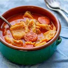 Un cuenco verde con callos a la madrileña, trozos de chorizo y un tenedor al fondo.