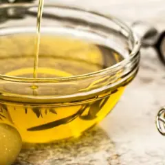 Un chorro de aceite de oliva crudo cae en un cuenco de cristal. Junto a él, aceitunas verdes y utensilios de cocina.