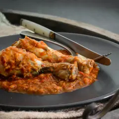 Dos muslos de pollo al chilindrón, servidos en un plato oscuro con cubiertos.