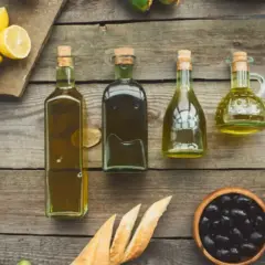 Botellas de aceite refinado de oliva junto a limones, hierbas frescas, pan y aceitunas sobre una mesa de madera.