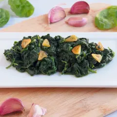Deliciosas espinacas salteadas con ajo laminado, servidas en un plato blanco. Acompañan ajos y hojas de espinaca fresca.
