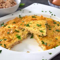 Tortilla de atún dorada y esponjosa, espolvoreada con perejil fresco. Un trozo cortado revela la textura jugosa.