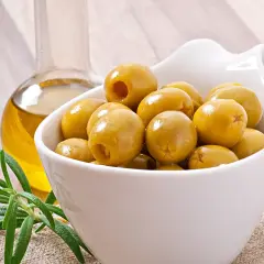 Un cuenco blanco lleno de olivas verdes, junto a una botella de aceite de oliva y una ramita de romero. Las olivas son saludables y deliciosas.