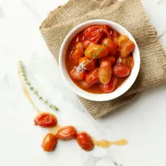 Un tazón blanco lleno de tomates cherry confitados, servido sobre arpillera, con algunos tomates sueltos y ramitas de tomillo en una superficie de mármol.