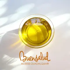 Aceite de oliva virgen extra en vaso de cristal, con reflejos de luz. Bajo, el logo "Buensalud Aceites Guadalquivir", indicando su bajo índice de acidez.
