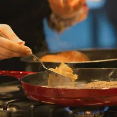 Una mano añade cebolla caramelizada a una sartén con una hamburguesa.