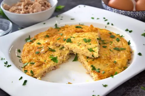 Tortilla de atún dorada y esponjosa, espolvoreada con perejil fresco. Un trozo cortado revela la textura jugosa.