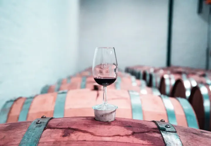 Copa de vino tinto sobre un barril de madera, con una bodega llena de barricas de vino crianza o reserva al fondo.