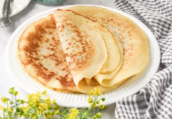 Deliciosos crepes sin gluten apilados en un plato blanco, con un toque de flores amarillas y una servilleta a cuadros.