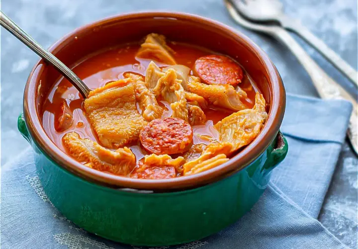 Un cuenco verde con callos a la madrileña, trozos de chorizo y un tenedor al fondo.