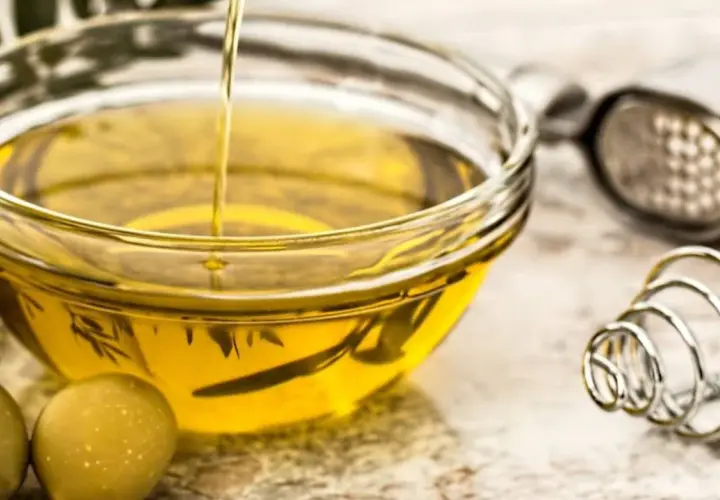 Un chorro de aceite de oliva crudo cae en un cuenco de cristal. Junto a él, aceitunas verdes y utensilios de cocina.