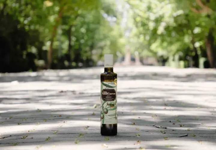 Aceite de oliva virgen extra de la almazara Pago de Valdecuevas, en botella oscura, sobre un camino de baldosas con árboles al fondo.