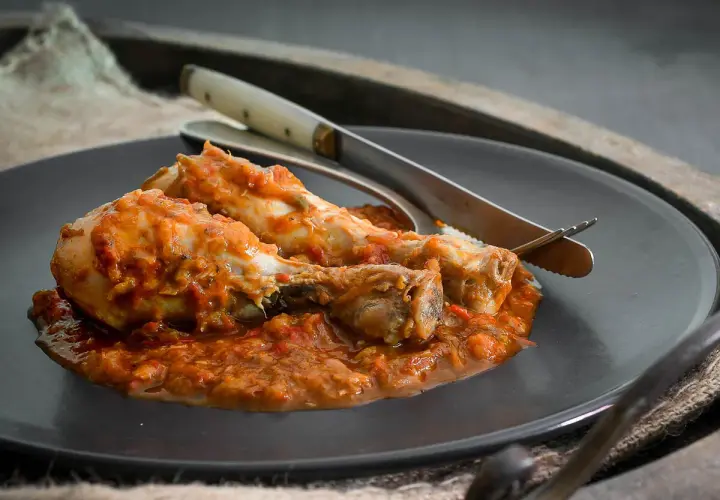 Dos muslos de pollo al chilindrón, servidos en un plato oscuro con cubiertos.