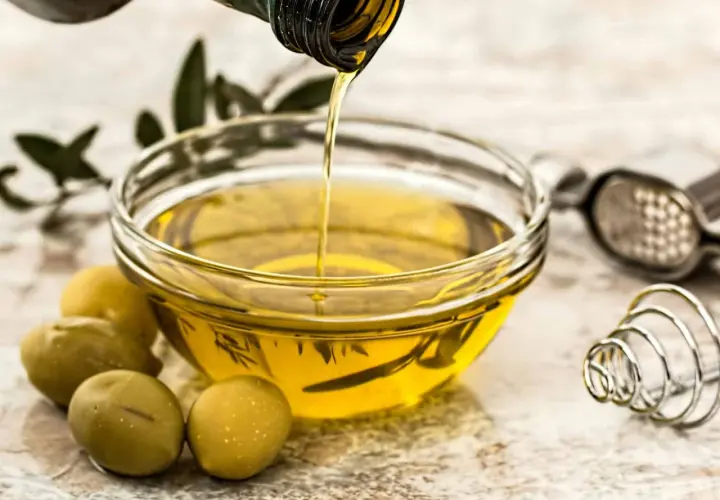 Aceite de oliva virgen extra vertiéndose en un cuenco, rodeado de aceitunas y hierbas. Un tesoro de vitaminas del aceite de oliva.