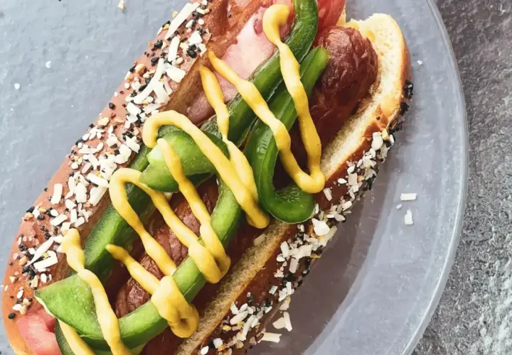 Un perrito caliente con pan cubierto de semillas, salchicha, pimientos verdes y mostaza amarilla.