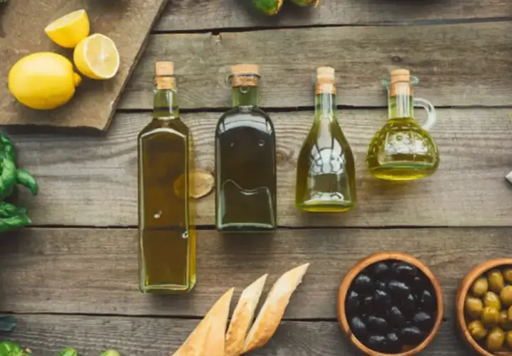 Botellas de aceite refinado de oliva junto a limones, hierbas frescas, pan y aceitunas sobre una mesa de madera.