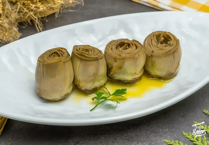 Cuatro alcachofas confitadas en aceite de oliva, servidas en plato blanco con perejil.