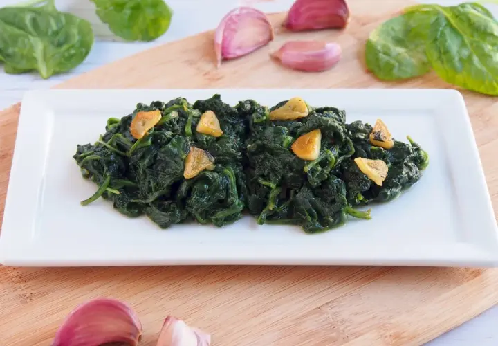 Deliciosas espinacas salteadas con ajo laminado, servidas en un plato blanco. Acompañan ajos y hojas de espinaca fresca.