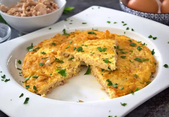 Tortilla de atún dorada y esponjosa, espolvoreada con perejil fresco. Un trozo cortado revela la textura jugosa.
