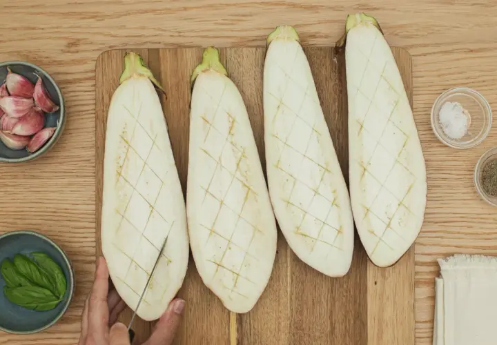 Berenjenas cortadas a la mitad, listas para ser rellenas. A su lado, ajos, albahaca, sal y pimienta.