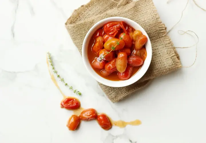 Un tazón blanco lleno de tomates cherry confitados, servido sobre arpillera, con algunos tomates sueltos y ramitas de tomillo en una superficie de mármol.