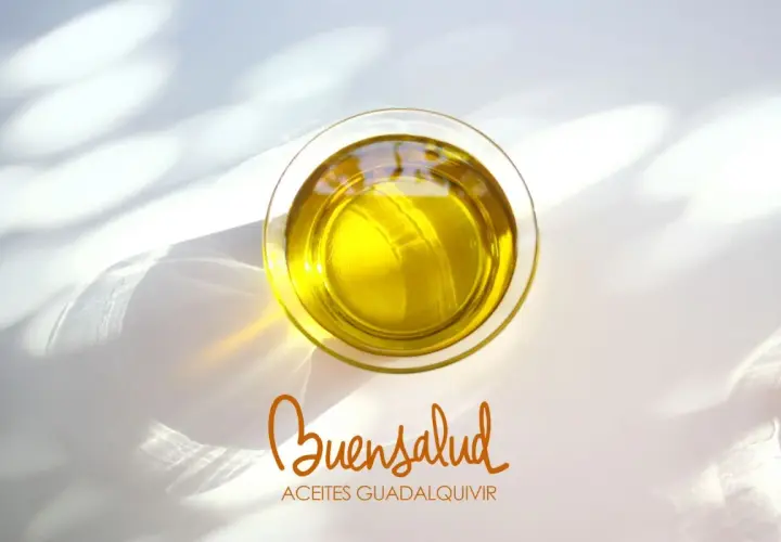 Aceite de oliva virgen extra en vaso de cristal, con reflejos de luz. Bajo, el logo "Buensalud Aceites Guadalquivir", indicando su bajo índice de acidez.