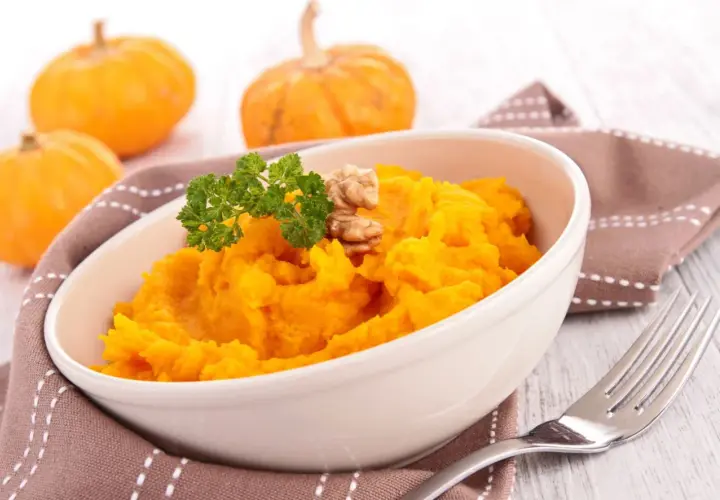 Un tazón de cremoso puré de calabaza, decorado con perejil y nueces, junto a calabazas pequeñas.