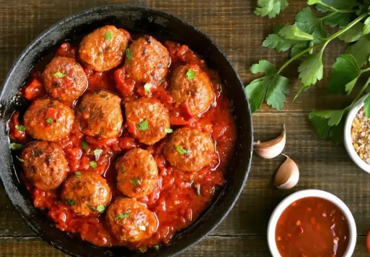Sabrosas albóndigas en salsa roja, servidas en una sartén de hierro fundido, con perejil fresco y especias al lado.