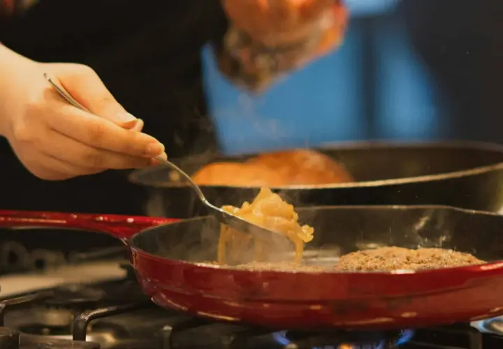 Una mano añade cebolla caramelizada a una sartén con una hamburguesa.