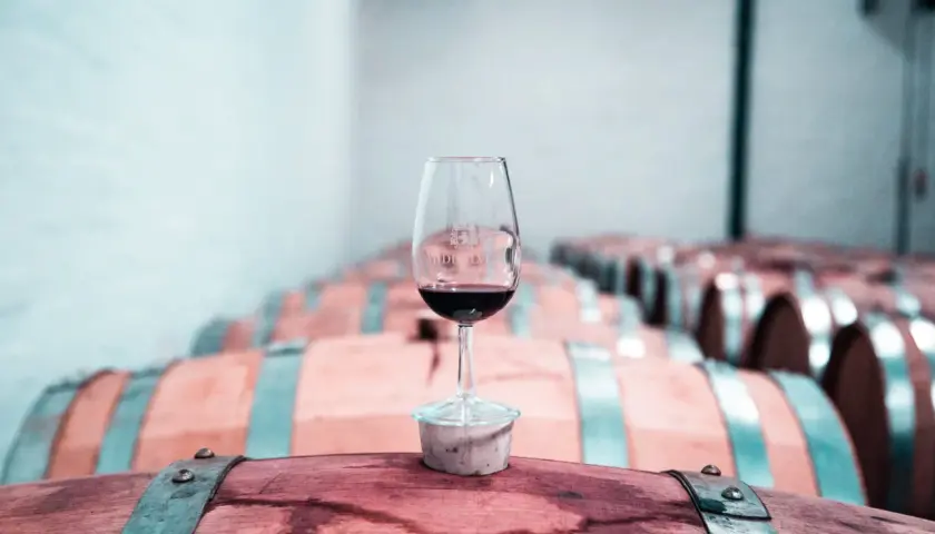 Copa de vino tinto sobre un barril de madera, con una bodega llena de barricas de vino crianza o reserva al fondo.