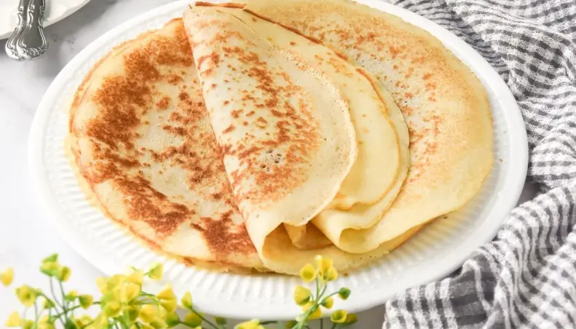 Deliciosos crepes sin gluten apilados en un plato blanco, con un toque de flores amarillas y una servilleta a cuadros.