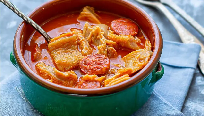 Un cuenco verde con callos a la madrileña, trozos de chorizo y un tenedor al fondo.