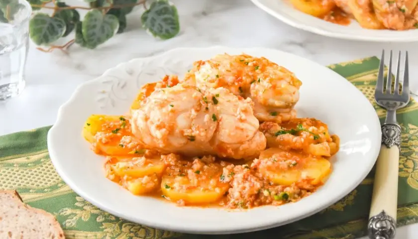 Rape con patatas en salsa de tomate, servido en plato blanco con tenedor y pan.