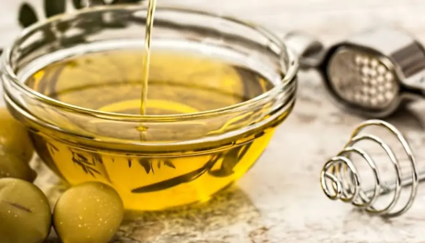 Un chorro de aceite de oliva crudo cae en un cuenco de cristal. Junto a él, aceitunas verdes y utensilios de cocina.