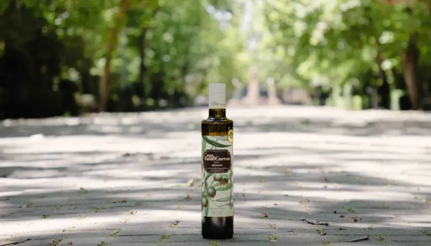 Aceite de oliva virgen extra de la almazara Pago de Valdecuevas, en botella oscura, sobre un camino de baldosas con árboles al fondo.