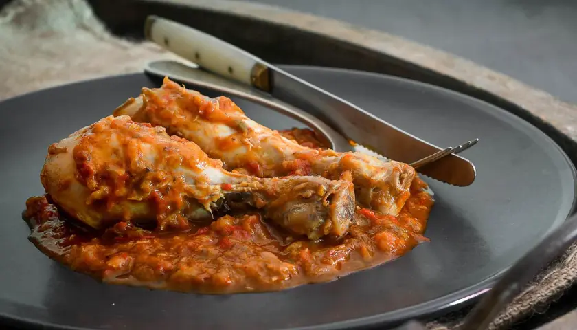 Dos muslos de pollo al chilindrón, servidos en un plato oscuro con cubiertos.