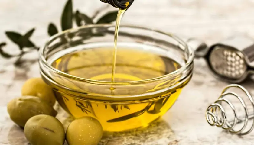 Aceite de oliva virgen extra vertiéndose en un cuenco, rodeado de aceitunas y hierbas. Un tesoro de vitaminas del aceite de oliva.