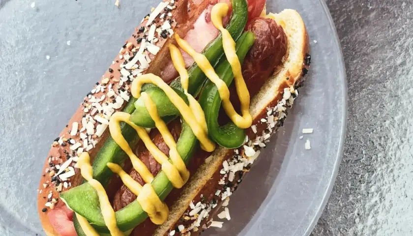 Un perrito caliente con pan cubierto de semillas, salchicha, pimientos verdes y mostaza amarilla.