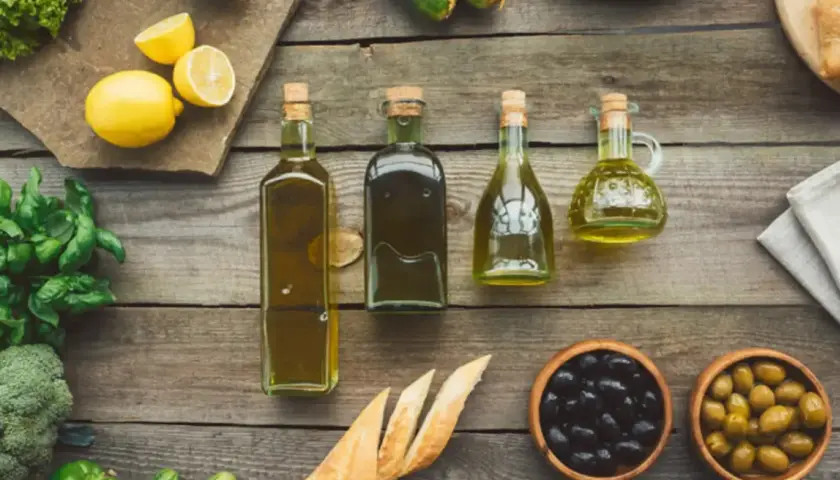Botellas de aceite refinado de oliva junto a limones, hierbas frescas, pan y aceitunas sobre una mesa de madera.