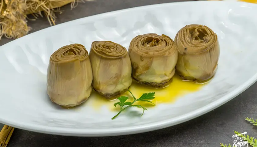 Cuatro alcachofas confitadas en aceite de oliva, servidas en plato blanco con perejil.