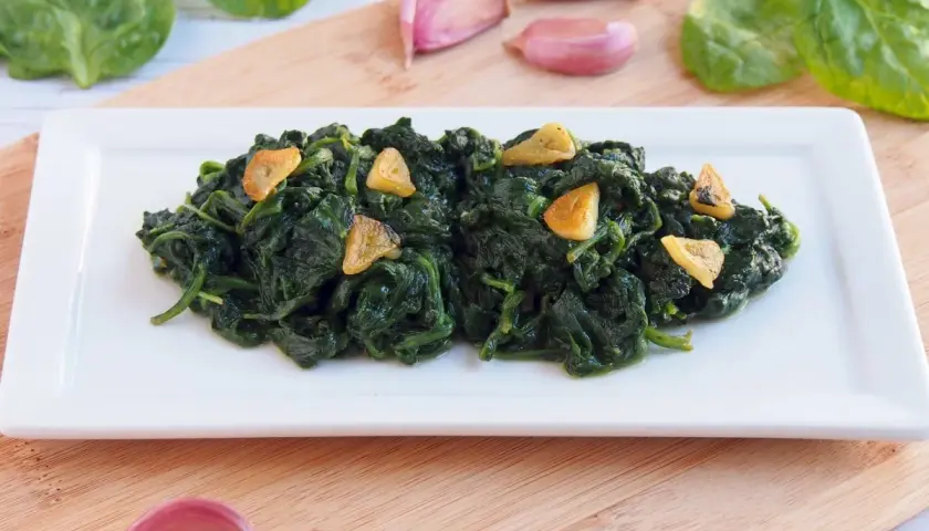 Deliciosas espinacas salteadas con ajo laminado, servidas en un plato blanco. Acompañan ajos y hojas de espinaca fresca.