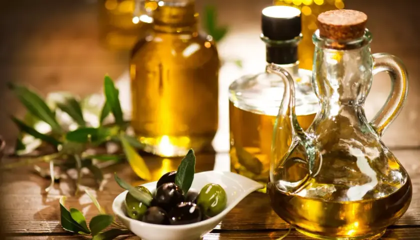 Aceite de oliva dorado en botellas y jarra, con aceitunas frescas y ramas de olivo. Descubre las propiedades del aceite de oliva.
