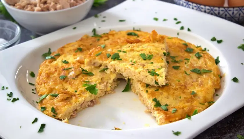 Tortilla de atún dorada y esponjosa, espolvoreada con perejil fresco. Un trozo cortado revela la textura jugosa.