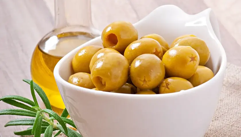 Un cuenco blanco lleno de olivas verdes, junto a una botella de aceite de oliva y una ramita de romero. Las olivas son saludables y deliciosas.