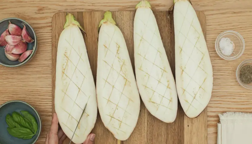 Berenjenas cortadas a la mitad, listas para ser rellenas. A su lado, ajos, albahaca, sal y pimienta.