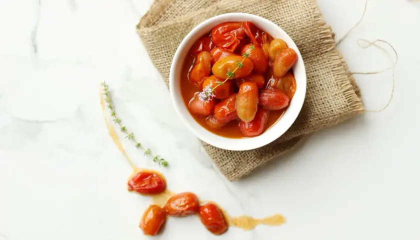 Un tazón blanco lleno de tomates cherry confitados, servido sobre arpillera, con algunos tomates sueltos y ramitas de tomillo en una superficie de mármol.