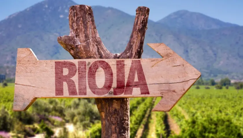 Flecha de madera rústica con la palabra "RIOJA" en rojo, apuntando hacia un paisaje de viñedos y montañas.