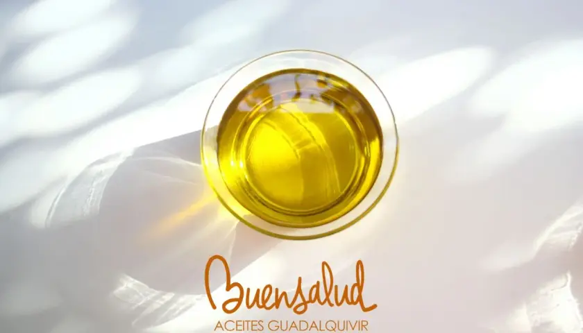 Aceite de oliva virgen extra en vaso de cristal, con reflejos de luz. Bajo, el logo "Buensalud Aceites Guadalquivir", indicando su bajo índice de acidez.