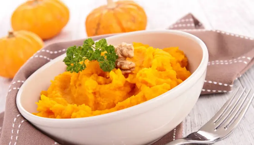 Un tazón de cremoso puré de calabaza, decorado con perejil y nueces, junto a calabazas pequeñas.